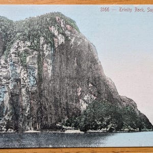 May include: Vintage postcard depicting Trinity Rock, Saguenay River. The image shows a large rock formation covered in trees, rising from the water. The sky is a pale blue, and the postcard has a slightly aged appearance.