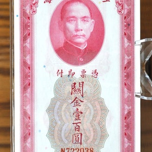 Puede incluir: Un billete de banco rosa y blanco con el texto "行銀央中" (Banco Central de China) y "壹百" (Cien) en caracteres chinos. El billete de banco presenta un retrato de un hombre con un traje rojo y un sello con el texto "關金壹百圓" (Sello de oro de cien yuanes). El billete de banco tiene el número de serie N722038 impreso en él.