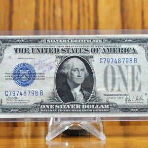 May include: A 1935 series United States one dollar silver certificate banknote featuring a portrait of George Washington, a blue treasury seal, and the serial number G79748798B, encased in a clear protective plastic holder on a display stand.