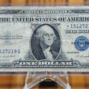 May include: A vintage one dollar silver certificate from the United States of America. The certificate features a portrait of George Washington and the text "This certifies that there is on deposit in the Treasury of the United States of America..."  The certificate is dated 1935.
