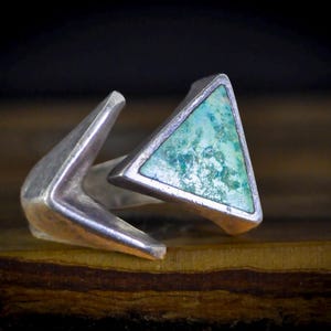 May include: A silver ring with a triangular turquoise stone. The ring has a unique design with two angular arms extending from the band, creating a modern and geometric look. The turquoise stone adds a pop of color.