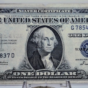 May include: A vintage one-dollar silver certificate from the United States, featuring a portrait of George Washington. The bill is in a protective case and displays text including "Silver Certificate" and "One Dollar."
