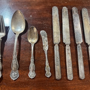 Puede incluir: Una colección de cubiertos de tono plateado que incluye un montón de tenedores, una cuchara grande, una cuchara pequeña, un cuchillo de untar pequeño y cuatro cuchillos de mesa. Cada pieza presenta un acabado desgastado y un patrón floral.