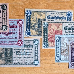May include: A selection of vintage German banknotes, with denominations ranging from 10 to 80. The notes display detailed illustrations of buildings and landscapes. The banknotes are in various colours, including pink, blue, and green, and are arranged on a wooden surface.