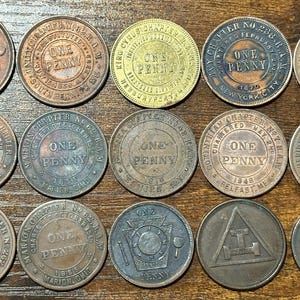 Puede incluir: Una colección de peniques antiguos de cobre, cada uno con diseños y textos intrincados. Las monedas varían en color, desde marrón claro hasta bronce oscuro, con inscripciones visibles como "ONE PENNY" y nombres de capítulos.