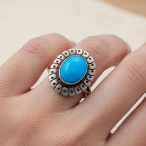 Puede incluir: Un anillo de plata con una piedra grande, ovalada, de color turquesa brillante. La piedra está engastada en un bisel decorativo y festoneado. El anillo está en un dedo, con un fondo neutro.