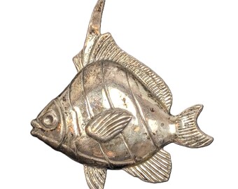 May include: A silver-coloured metal fish brooch. The brooch has a detailed design with fins, scales, and a prominent dorsal fin. The fish is facing left, with its mouth slightly open. The brooch has a vintage or antique appearance.