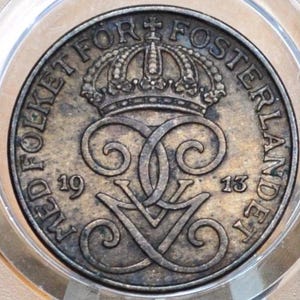 Könnte beinhalten: Eine bronzene Münze mit der Aufschrift "Med Folket For Fosterlandet" und dem Jahr "1913" am Rand. In der Mitte der Münze befindet sich eine Krone über einem stilisierten "V" mit einem dekorativen Design.