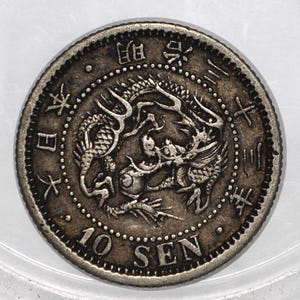 May include: A circular silver-toned Japanese 10 Sen coin featuring a central embossed dragon design. The coin displays Japanese characters along the upper rim and the text 10 SEN at the bottom, with a beaded border encircling the entire edge.