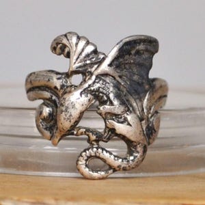 May include: A silver dragon ring with detailed features. The dragon has wings and a curled tail. The ring is displayed on a clear glass surface, with a wooden background.