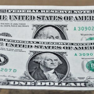 May include: Two U.S. one-dollar bills, slightly curved, displaying the portrait of George Washington. The bills feature the text "THE UNITED STATES OF AMERICA" and the number "1" in multiple locations. The bills are on a wooden surface.
