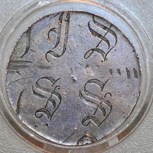 Può includere: Un oggetto metallico circolare e invecchiato con quattro disegni stilizzati della lettera "S". Le lettere sono in una scrittura decorativa e l'oggetto è racchiuso in una cornice circolare trasparente. Il metallo ha un aspetto usurato e strutturato.