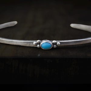 May include: A silver cuff bracelet with a central oval turquoise stone and small beaded accents. The bracelet has a simple, elegant design, perfect for everyday wear. The turquoise stone is a vibrant blue color.