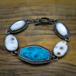 Può includere: Bracciale color argento con una grande pietra turchese e pietre ovali più piccole bianche e marroni. Il bracciale ha un design a catena e una chiusura a fermaglio. Le pietre sono incastonate in un bordo decorativo a forma di corda.