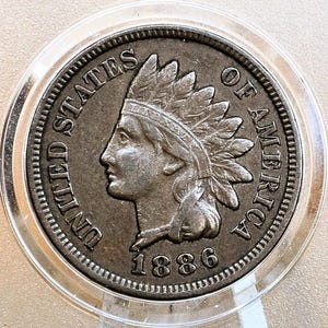 May include: A copper-colored 1886 United States Indian Head penny coin. The coin features a profile of a Native American woman wearing a feathered headdress. The inscription "UNITED STATES OF AMERICA" is around the rim of the coin.