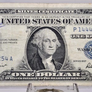 May include: A vintage one-dollar silver certificate from the United States, featuring a portrait of George Washington. The bill has the text "ONE DOLLAR" and "SILVER CERTIFICATE". The bill is in a protective case.