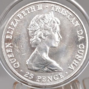 Puede incluir: Una moneda de plata de 25 peniques con el perfil de la reina Isabel II. La moneda está inscrita con "QUEEN ELIZABETH II TRISTAN DA CUNHA". La moneda está dentro de un soporte protector transparente.