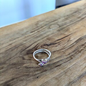 May include: A silver ring with a pink stone design. The ring is on a wooden surface.