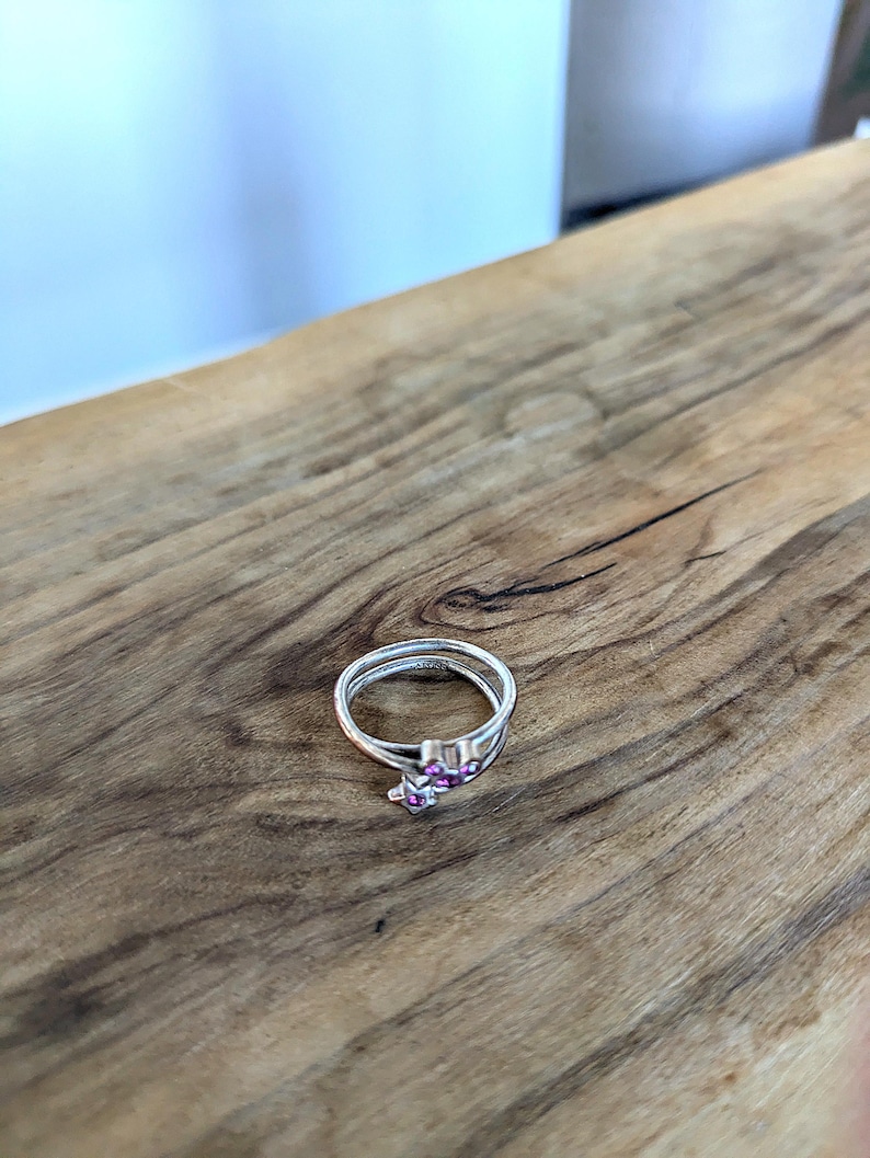May include: A silver ring with a pink stone design. The ring is on a wooden surface.