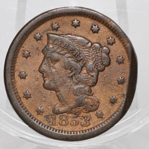 Pode incluir: Uma moeda cor de cobre com o perfil da Liberdade usando um diadema com a palavra LIBERTY. A data 1853 está gravada na parte inferior, rodeada por treze estrelas de cinco pontas dispostas em círculo ao longo da borda externa.