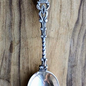 May include: Silver souvenir spoon with a detailed figure of a man and the word "HEIDELBERG" at the top. The spoon has an ornate handle with a twisted design and a round bowl.