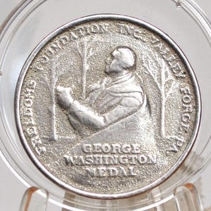 Pode incluir: Uma medalha de prata de George Washington dentro de uma caixa de plástico transparente. A medalha apresenta um relevo de George Washington e o texto "GEORGE WASHINGTON MEDAL". As palavras "FREEDOM FOUNDATION INC. VALLEY FORGE" estão inscritas na borda.