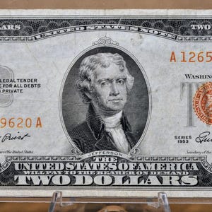 May include: A vintage two-dollar bill featuring a portrait of Thomas Jefferson. The bill has a red seal and serial numbers, with the text "The United States of America" and "Two Dollars" prominently displayed. The bill is encased in a protective sleeve.