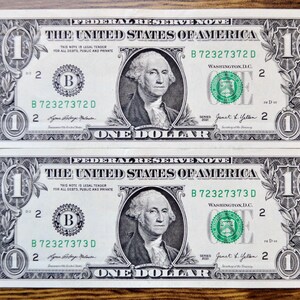 May include: Two United States one-dollar bills featuring a portrait of George Washington in the center. The bills display the text Federal Reserve Note, The United States of America, One Dollar, and serial numbers B 72327372 D and B 72327373 D.