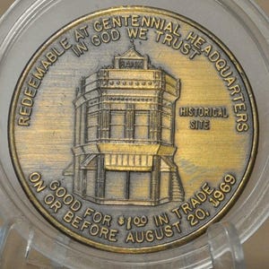 May include: A gold-toned commemorative coin encased in clear plastic. The coin features a detailed illustration of a building labeled "BANK" and "HISTORICAL SITE". Text around the edge reads "REDEEMABLE AT CENTENNIAL HEADQUARTERS IN GOD WE TRUST" and "GOOD FOR $1.00 IN TRADE ON OR BEFORE AUGUST 20, 1969".
