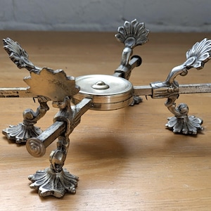 May include: A silver-toned metal trivet with ornate, floral-inspired details. The trivet has a circular center and four arms extending outwards, each supported by decorative legs. The arms have decorative finials at the ends.
