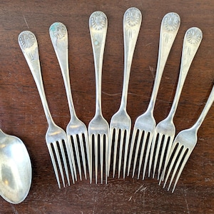 Puede incluir: Un conjunto de cubiertos antiguos de plata, que incluye una cuchara y siete tenedores. Los utensilios están dispuestos sobre una superficie de madera oscura. Cada pieza tiene un diseño decorativo en el mango.