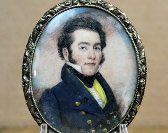 May include: Oval portrait of a man in a dark blue coat with gold buttons, a yellow vest, and a white shirt. The portrait is set in an ornate silver-toned frame and displayed on a clear stand. The man has dark hair and blue eyes.