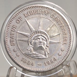 May include: A silver coin commemorating the Statue of Liberty Centennial. The coin features the statue's face at the center, surrounded by the text "STATUE OF LIBERTY CENTENNIAL" and the years "1884 - 1984". The coin is encased in a clear protective holder.