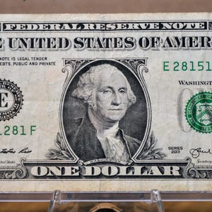 Puede incluir: Un billete de un dólar estadounidense con un retrato de George Washington en el centro. El billete incluye el texto THE UNITED STATES OF AMERICA, FEDERAL RESERVE NOTE y el número de serie E 28151281F impreso en tinta verde.
