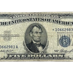 May include: A vintage 1953 five-dollar silver certificate banknote featuring a portrait of Abraham Lincoln in the center. The bill displays the text The United States of America, Five Dollars, and serial number 10662983 A with star symbols.
