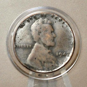 May include: A 1947 Lincoln wheat penny coin in a protective plastic holder. The coin features a profile of Abraham Lincoln, the word "LIBERTY", and the year 1947. The coin is encased in a clear, circular holder.