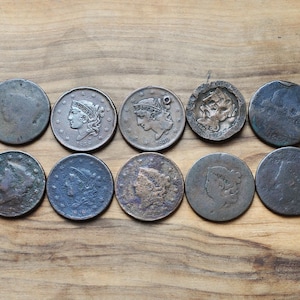 May include: A collection of ten antique coins, arranged in two rows on a wooden surface. The coins vary in size and condition, with visible details of portraits and inscriptions. The coins are in shades of silver, bronze, and dark grey.