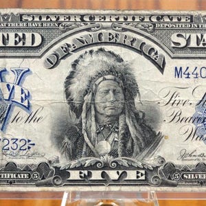 Peut inclure: Un ancien billet de cinq dollars en argent des États-Unis. Le billet présente un portrait d'un Amérindien coiffé d'une coiffe. Le texte comprend "Silver Certificate" et "Five Dollars". Le billet est en nuances de bleu et de gris.