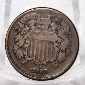 May include: A round copper-coloured two-cent coin featuring a central shield design with vertical stripes, surrounded by laurel wreaths. The text IN GOD WE TRUST is inscribed on a ribbon above the shield, and the year 1865 is printed at the bottom.