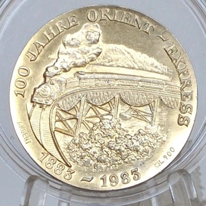 Puede incluir: Una moneda conmemorativa dorada que celebra el 100 aniversario del Orient Express. La moneda presenta un relieve detallado de un tren de vapor sobre un puente, con el texto "100 Jahre Orient Express" y los años 1883-1983.