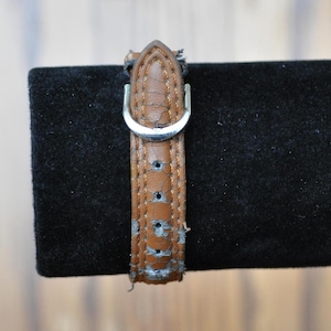 May include: A brown leather watch strap with a silver buckle. The strap has a worn look and is showing signs of age.