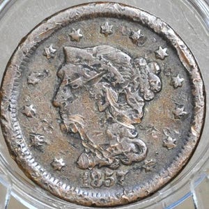 Puede incluir: Primer plano de una moneda antigua dentro de un soporte de plástico transparente. La moneda presenta el perfil de una mujer, rodeada de estrellas, con el año 1857. La moneda tiene un aspecto desgastado y envejecido.