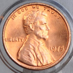 May include: A copper-colored 1975 Lincoln penny, encased in a clear plastic holder. The obverse features a profile of Abraham Lincoln, with the words "LIBERTY" and "IN GOD WE TRUST".