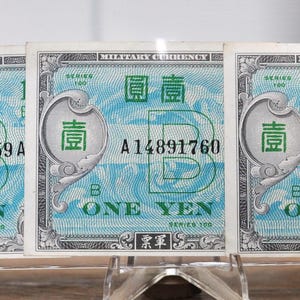 May include: Three vintage One Yen military currency notes. Each note has a light blue and white design with Chinese characters and the text "ONE YEN". The notes are displayed in a clear protective sleeve.