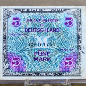 Può includere: Una banconota vintage da cinque marchi dell'Autorità Militare Alleata in Germania, con il testo "Umlauf Gesetzt in Deutschland" e il numero di serie 008361754. La banconota ha un design blu e viola con il numero 5 in ogni angolo.