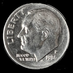 May include: A silver US dime coin with the profile of President Franklin D. Roosevelt on the obverse. The coin has the inscription "LIBERTY" on the left side and "IN GOD WE TRUST" and "1986" on the bottom.