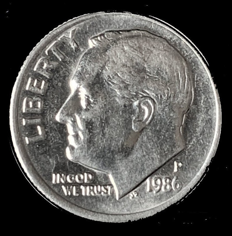 May include: A silver US dime coin with the profile of President Franklin D. Roosevelt on the obverse. The coin has the inscription "LIBERTY" on the left side and "IN GOD WE TRUST" and "1986" on the bottom.