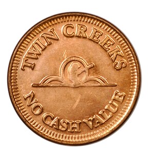 May include: A round copper-colored metal token featuring the embossed text TWIN CREEKS at the top and NO CASH VALUE at the bottom. In the center, there is a circular emblem depicting a stylized figure holding a long object above a horizontal line.