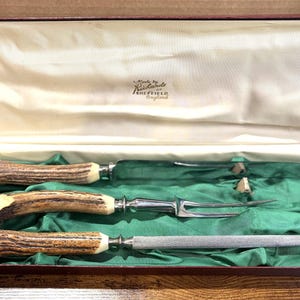 May include: A vintage carving set presented in a box. The set includes a carving knife, fork, and sharpening steel. The handles are made of a brown and cream-coloured material, and the blades are silver. The box is lined with green fabric.