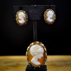 May include: A white ceramic mug with a black handle and the text "But First, Coffee" in a bold, black font. The mug is set against a blurred background. The mug appears to be approximately 10 cm tall.